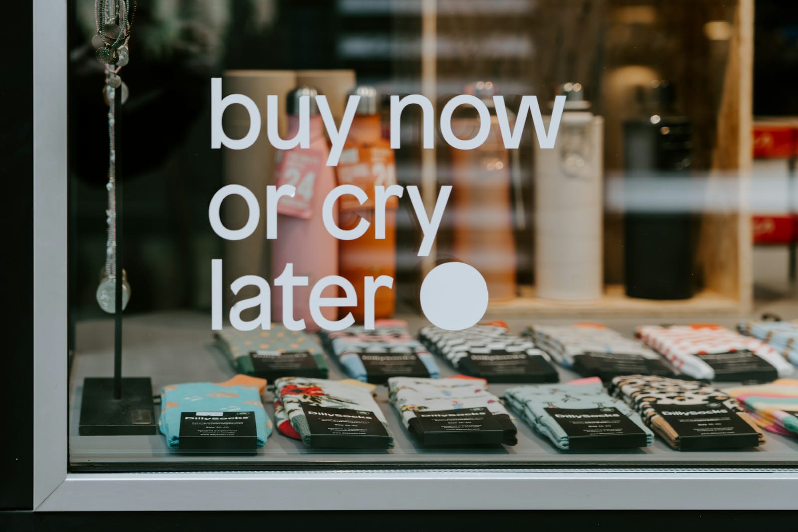Witte raamsticker met de tekst 'buy now or cry later' op een etalageruit met sokken erachter